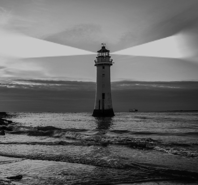 Fotografia in bianco e nero di un faro solitario su scogli, mare mosso e cielo nuvoloso